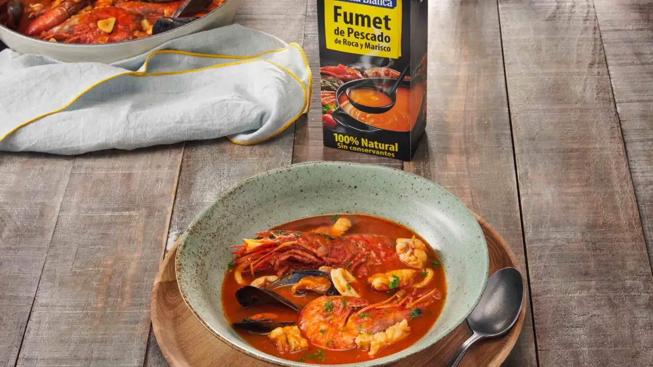 Un plato humeante de zarzuela de pescado, con gambas, mejillones y calamares, junto a un cartón de fumet de pescado.