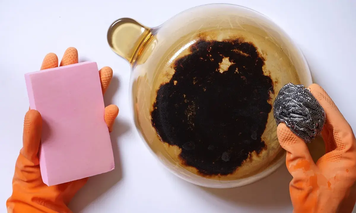Manos con guantes naranjas sostienen una esponja rosa y un estropajo metálico, listos para aprender como quitar el quemado de una olla.