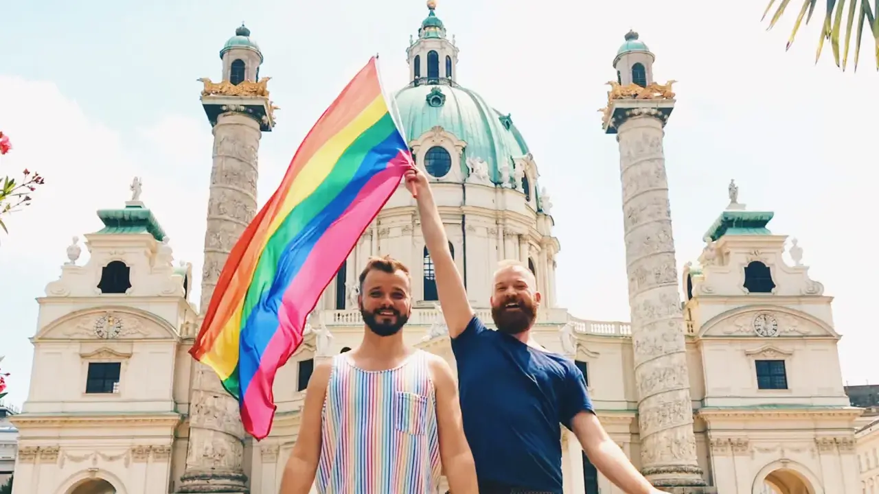 Two men proudly hold a rainbow flag in front of a grand building in Austria, celebrating gay pride.