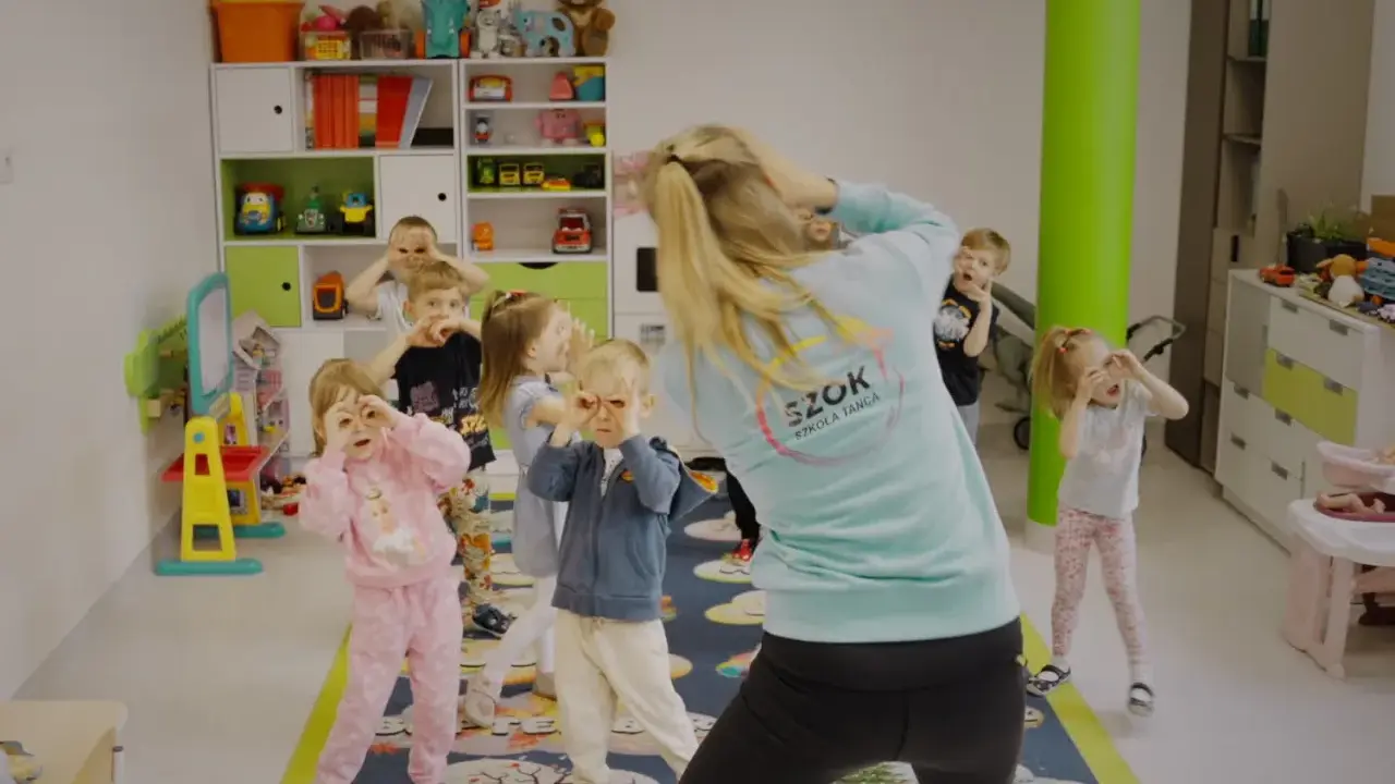Dzieci na zajęciach tańca wykonują proste układy taneczne do znanych piosenek, naśladując ruchy nauczycielki.