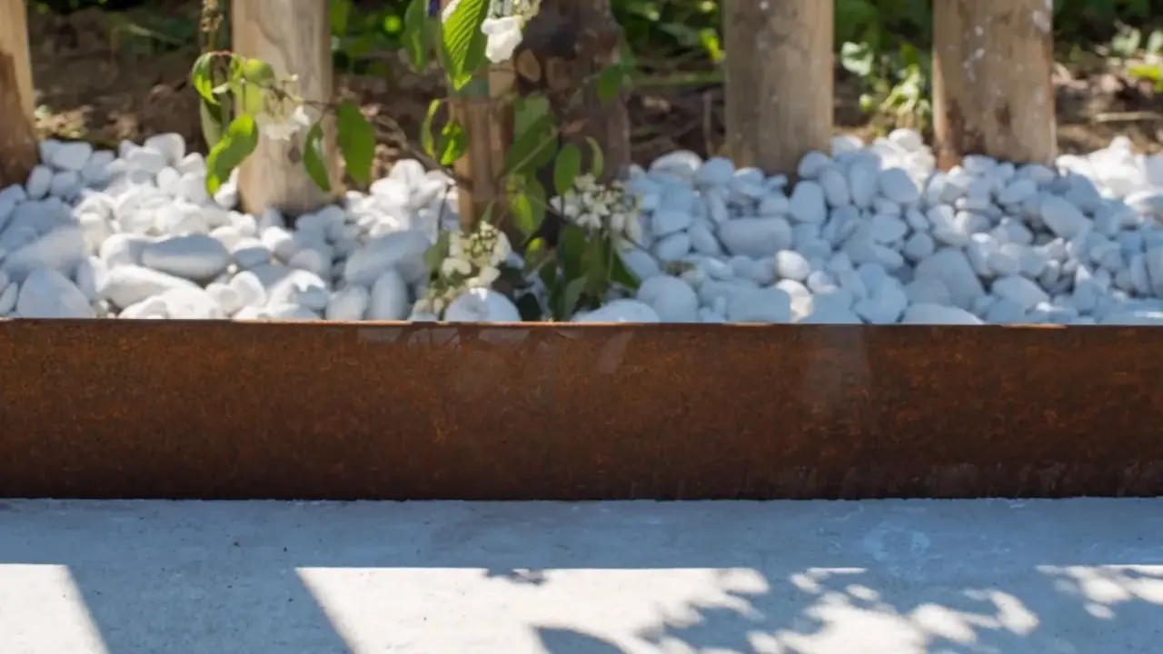 Parterre devant maison sans entretien, avec des galets blancs, des piquets en bois et une bordure rouillée. Des fleurs blanches éclosent.
