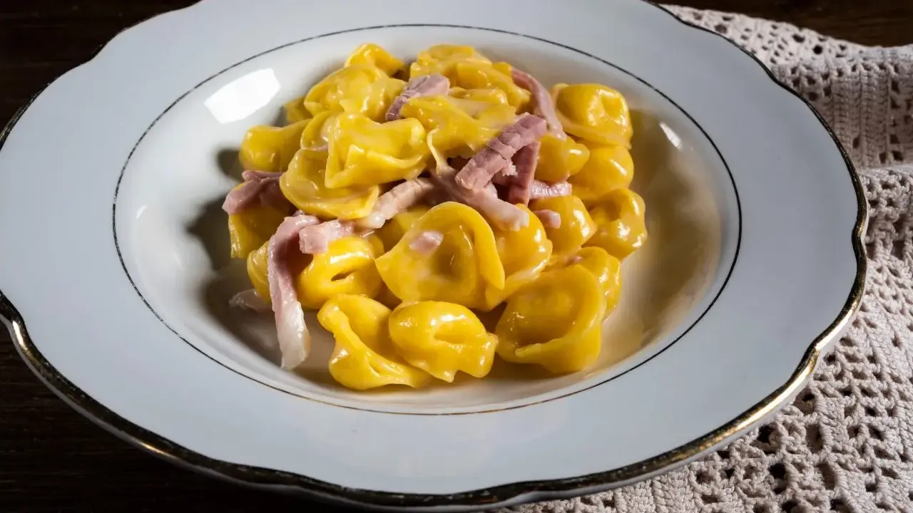 Tortelli ripieni in salsa cremosa con prosciutto, un classico tra i piatti tipici di Parma, serviti in un piatto bianco bordato d'oro.