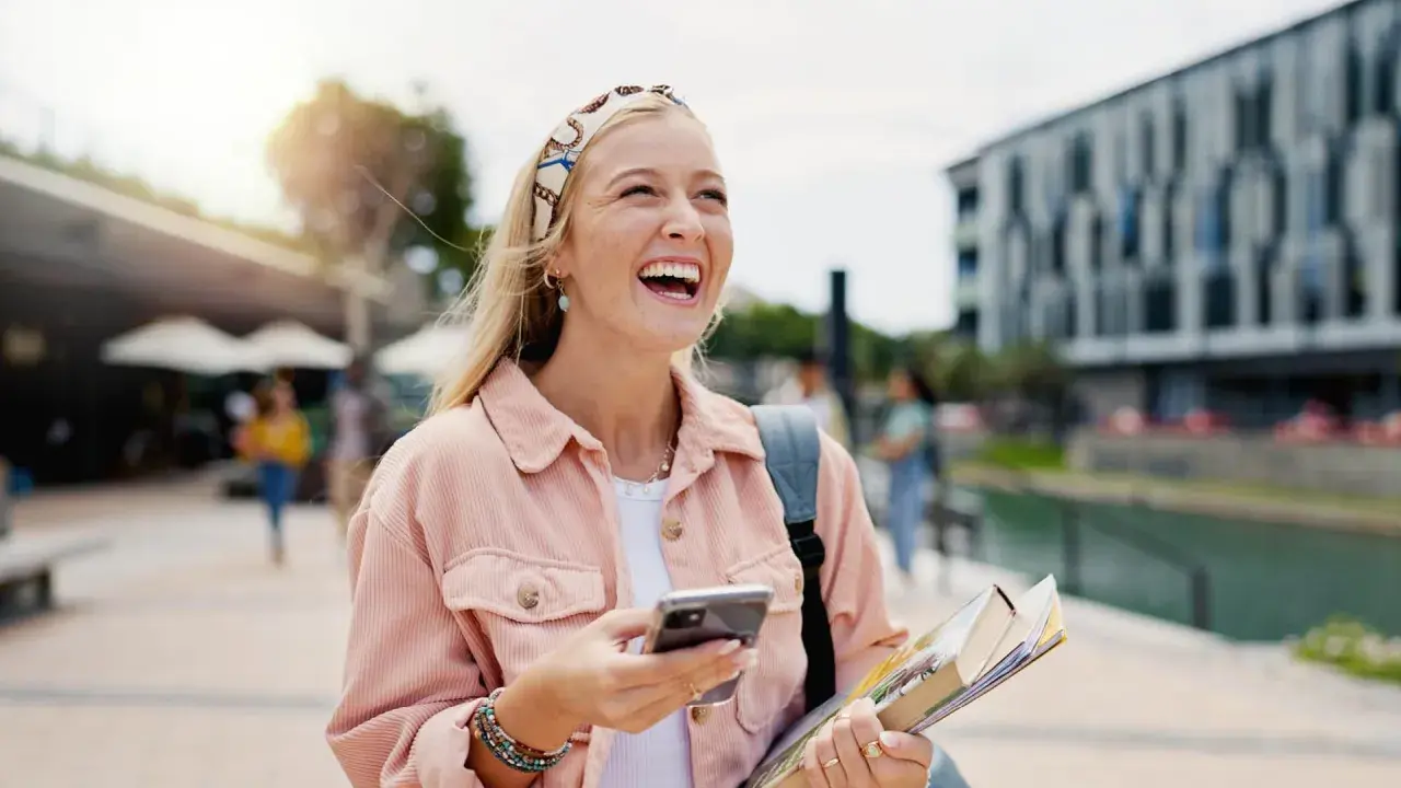 Szczęśliwa studentka z polską maturą, która planuje studia za granicą, śmieje się, trzymając telefon i książki.