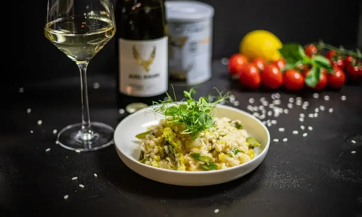 Aksamitne risotto z kurczakiem i szparagami, udekorowane kiełkami. Obok kieliszek białego wina i butelka.