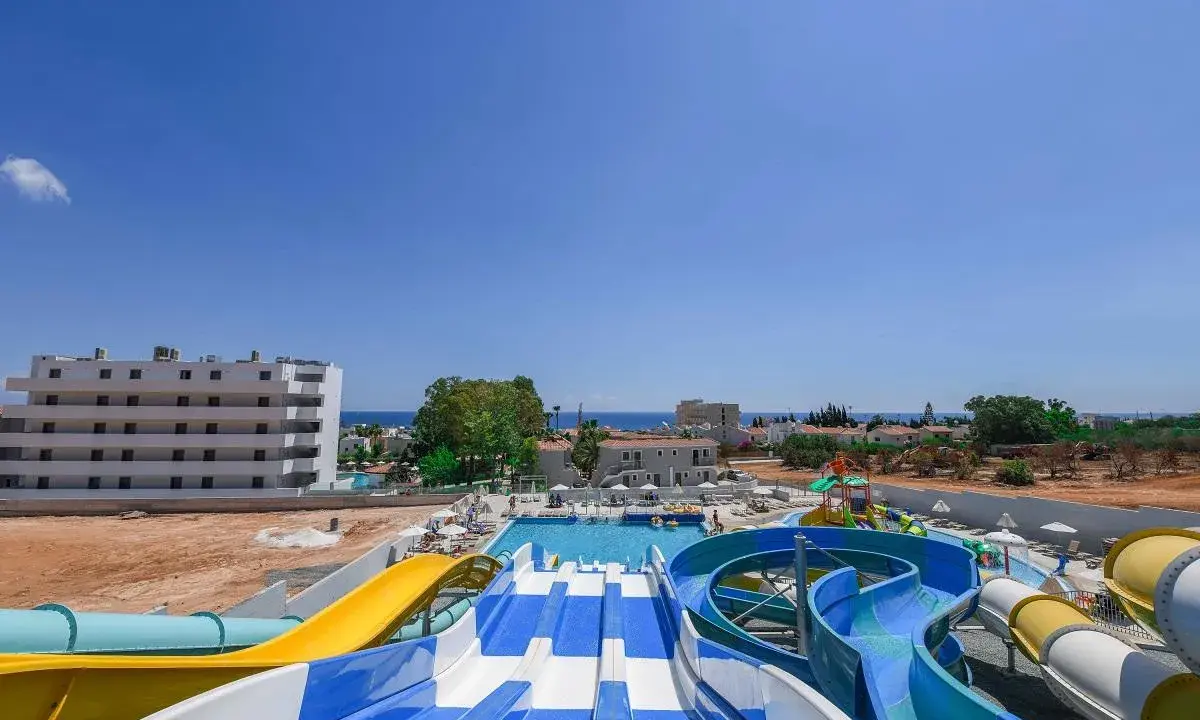 Kolorowe zjeżdżalnie wodne i baseny w Narcissos Waterpark Resort, z hotelami i morzem w tle.