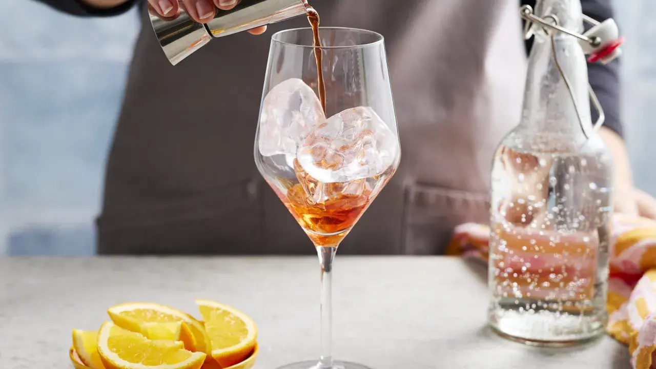 Ein Aperol-Spritz-Cocktail mit Prosecco wird zubereitet. Eiswürfel im Glas, daneben Orangenstücke und eine Flasche Sprudelwasser.