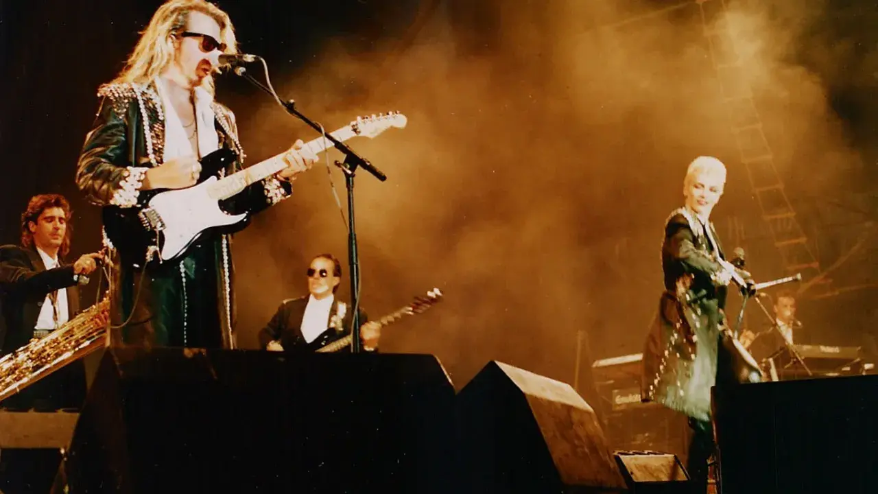 Medlemmar av Eurythmics på scen. Dave Stewart spelar gitarr, Annie Lennox sjunger. En saxofonist och en keyboardist syns också.