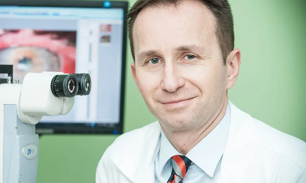 Okulista z uśmiechem przy aparaturze do badań. Na ekranie widok oka, sugerujący diagnostykę przed przeszczepem rogowki.