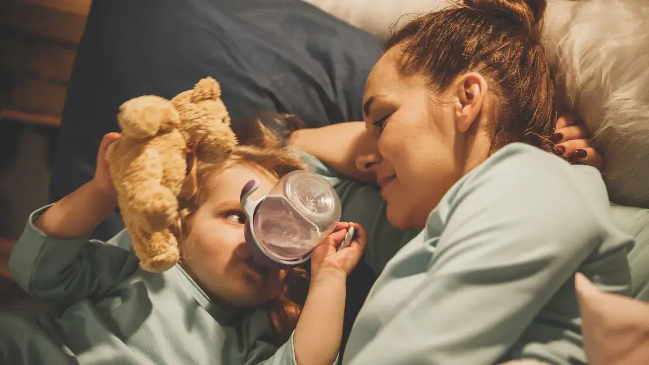 Mama opiekuje się dzieckiem z rotawirusem. Podaje mu wodę z kubeczka, a obok leży pluszowy miś.