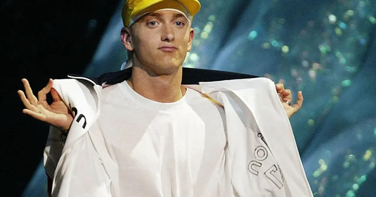 Slim Shady w żółtej czapce i białej bluzie, z gestem dłoni.