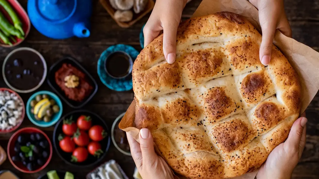 Hände halten ein frisch gebackenes Ramadan Brot, bestreut mit Sesam und Schwarzkümmel. Daneben liegen kleine Schalen mit Oliven, Tomaten und anderen Köstlichkeiten.