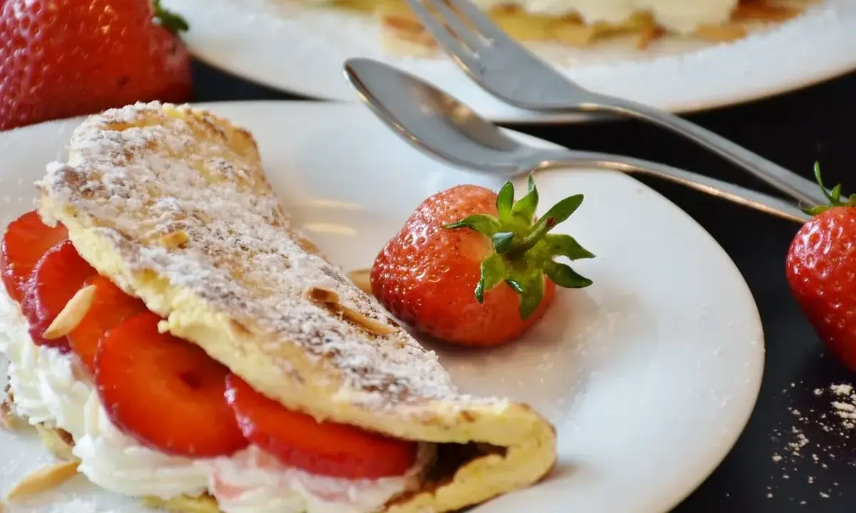 Puszysty omlet z białek, nadziany kremem i truskawkami, posypany cukrem pudrem. Obok leżą świeże owoce i sztućce.