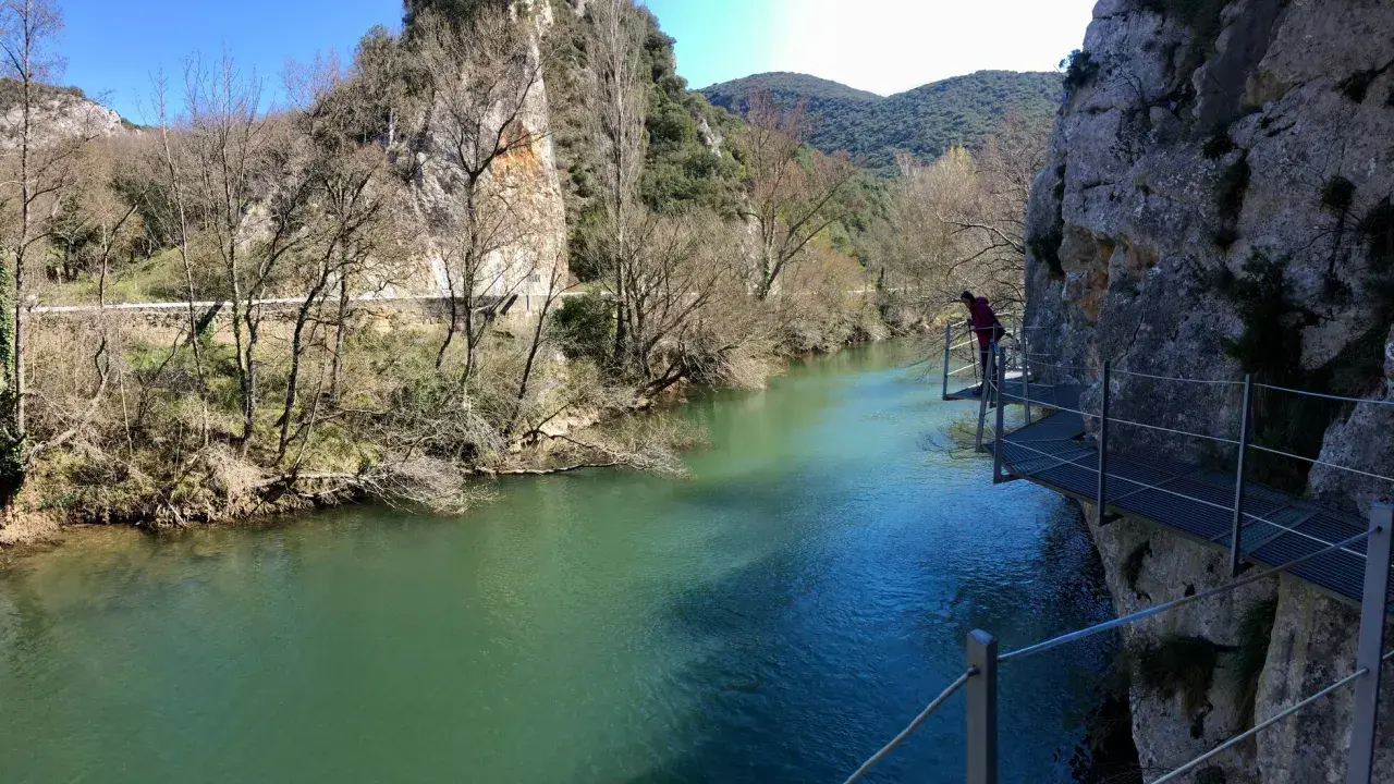 Persona camina por pasarela metálica junto a río turquesa, explorando las rutas de Burgos. Paisaje rocoso y vegetación de ribera.
