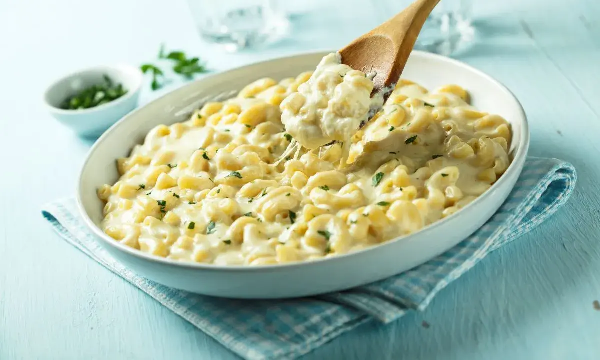Pyszne, kremowe mac n cheese z ziołami, podane w białej misce. Drewniana łyżka nabiera porcję ciągnącego się sera.