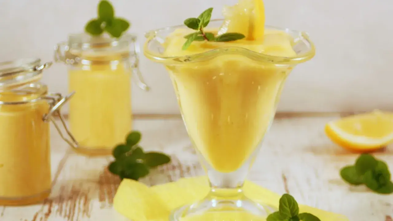 Pyszny lemon curd przepis w pucharku, ozdobiony cytryną i miętą. Obok słoiczki z kremem.