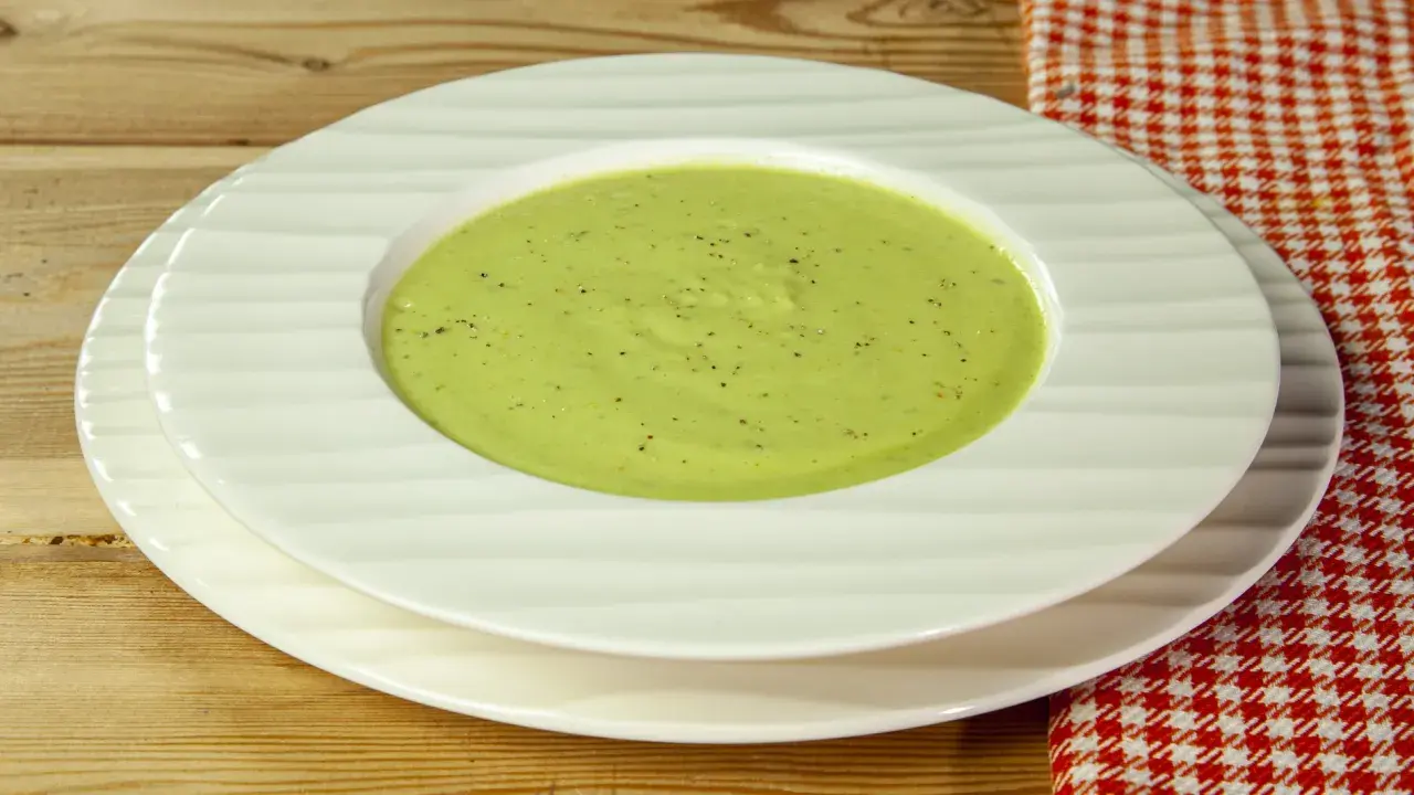 Una ciotola di cremosa crema di zucchine, condita con pepe nero, servita su piatti bianchi su un tavolo di legno.