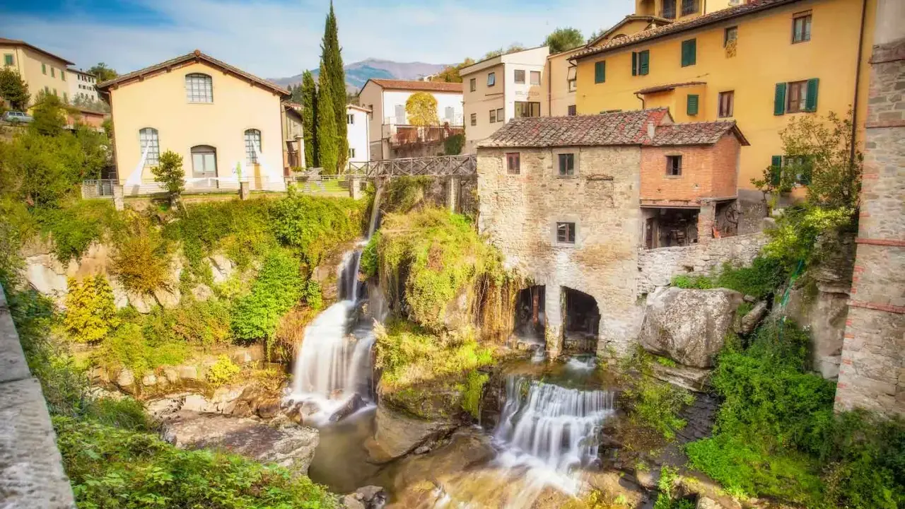 Cascata e mulino in pietra, tra i borghi della Toscana. Case gialle e verdi colline sullo sfondo.