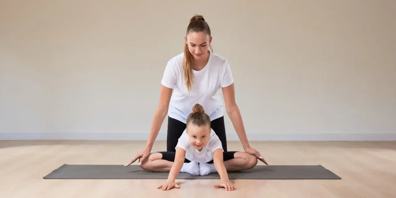 Frau und Kind machen Yoga. Das Kind krabbelt auf der Matte, die Frau unterstützt es. Yoga mit Kindern unter 3 Jahren fördert spielerisch die Bewegung.