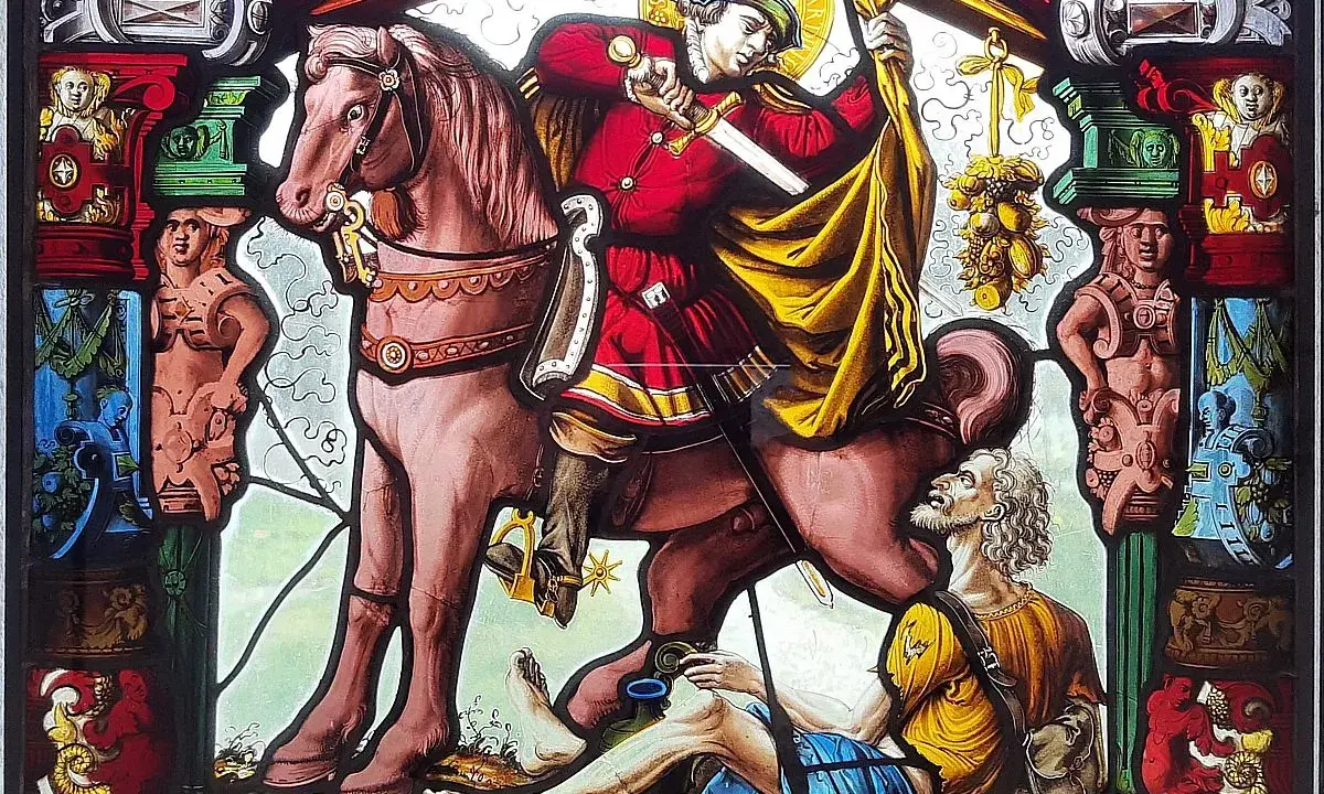 Święty Gracja na koniu przecina płaszcz dla żebraka. Witraż pełen symboli i postaci.