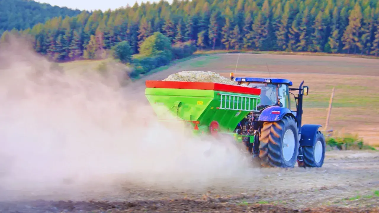 Traktor rozsiewa wapno do ziemi, tworząc chmurę pyłu na polu. W tle las i wzgórza.