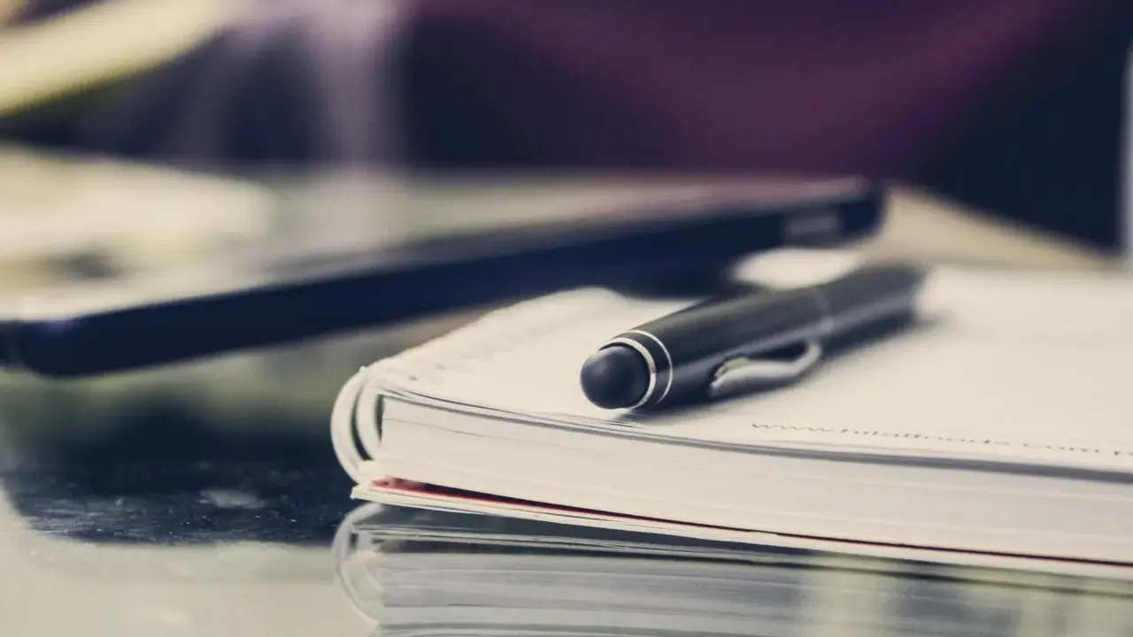 Czarny długopis spoczywa na otwartym notatniku. Obok leży laptop, gotowy do pracy. FGŚP.