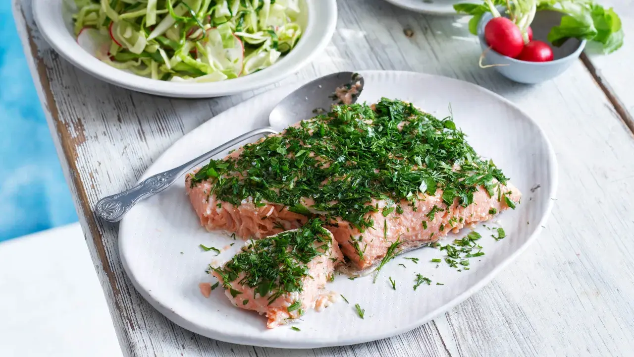 En saftig bit confiterad lax, generöst toppad med färska örter, serveras på en vit tallrik. Bredvid syns en fräsch sallad och några röda rädisor.