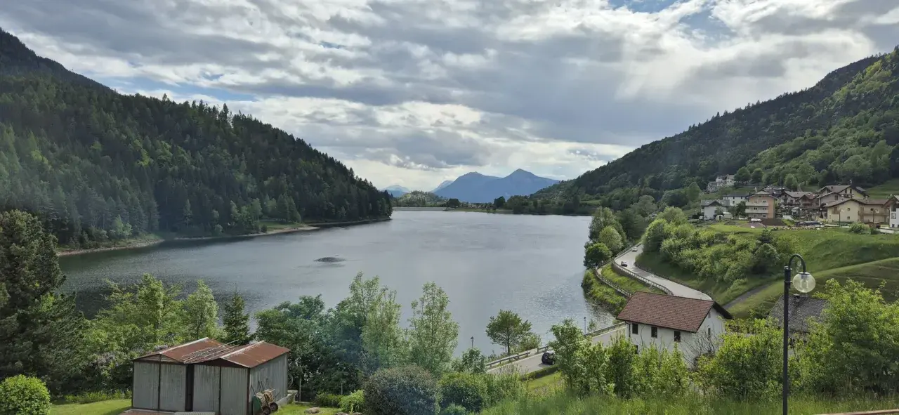 Panoramica del lago di Stramentizzo circondato da montagne verdi e un villaggio pittoresco. Nuvole minacciose nel cielo.