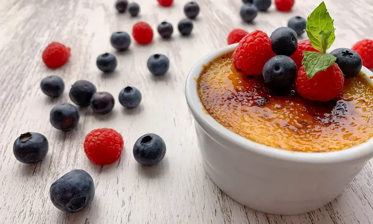 Creme brulee mit karamellisierter Kruste, garniert mit frischen Himbeeren und Blaubeeren, umgeben von weiteren Beeren auf hellem Holz.
