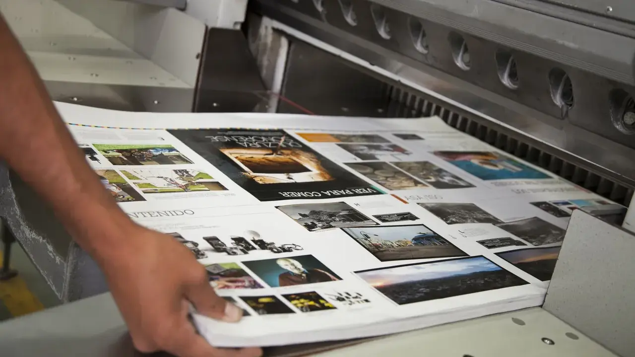 Mani che sistemano fogli stampati su una macchina da taglio, parte della post produzione di una rivista.