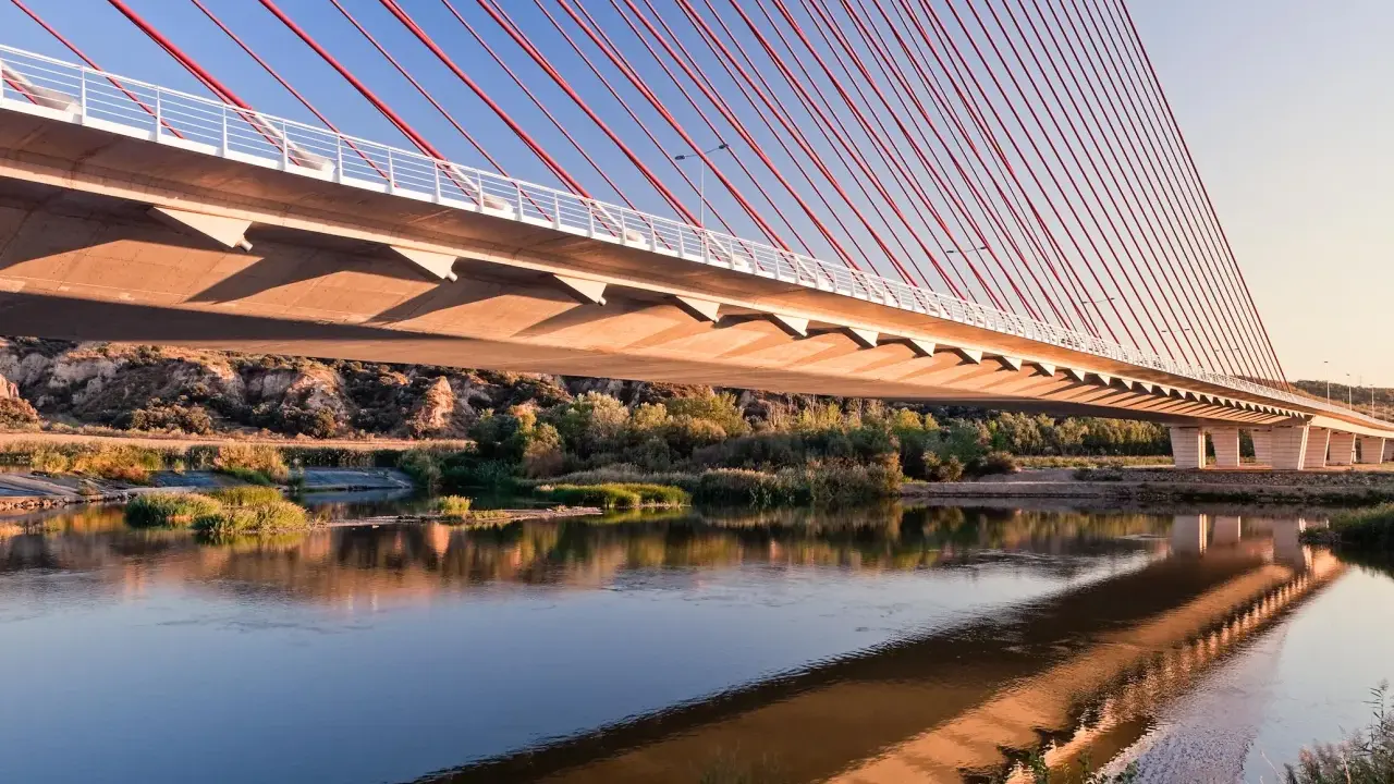 El puente de Castilla-La Mancha se refleja en el río, con sus cables rojos y el cielo azul.