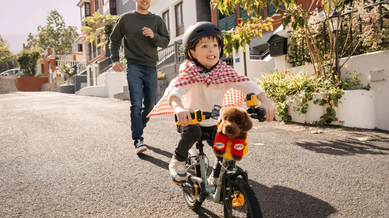 Un bambino con un mantello da supereroe prova ad andare in bicicletta, mentre suo padre corre dietro per aiutarlo. Mio figlio non riesce a pedalare, ma si diverte un mondo!