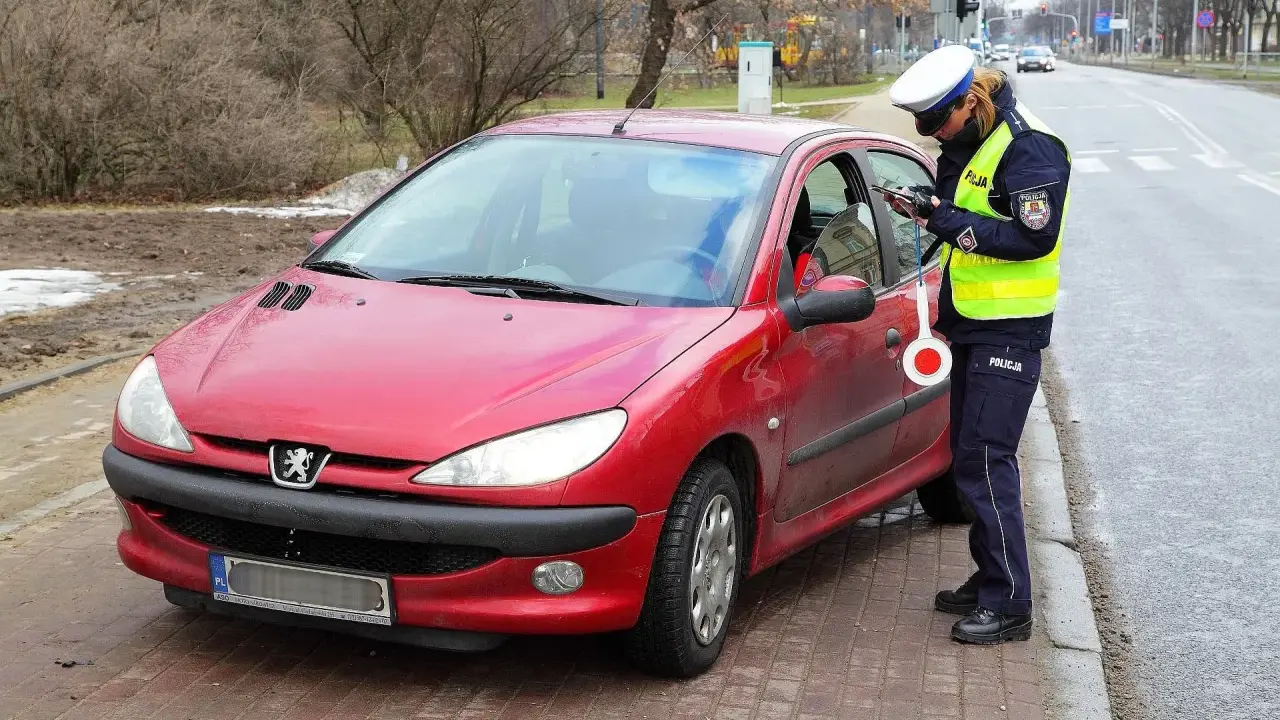 Policjantka w kamizelce odblaskowej i czapce spisuje kierowcę przy czerwonym samochodzie.
