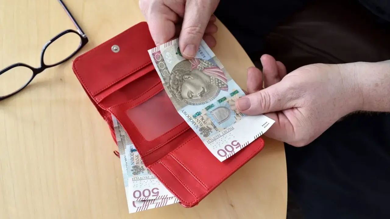 Ręce wkładają banknot 500 zł do czerwonego portfela. Obok okulary. Może to być pomocne przy sprawdzaniu 14 emerytury tabeli netto.