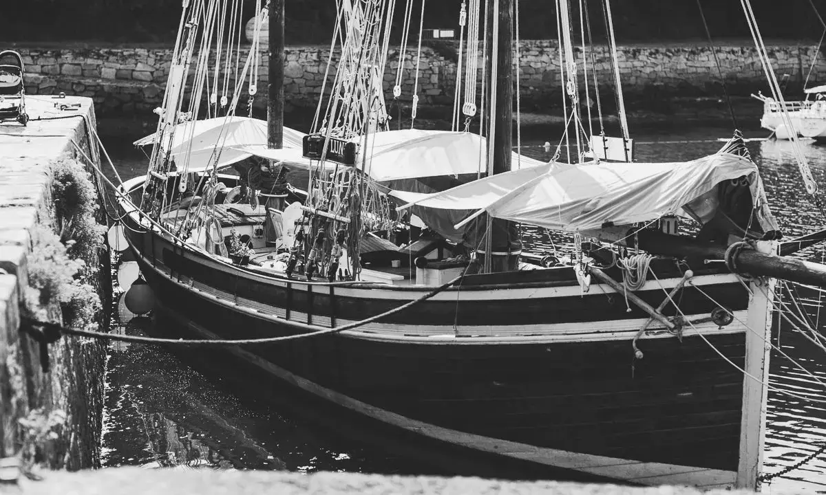 Bateau amarré, voiles repliées. On aperçoit la proue, le babord et le tribord. La poupe est moins visible.