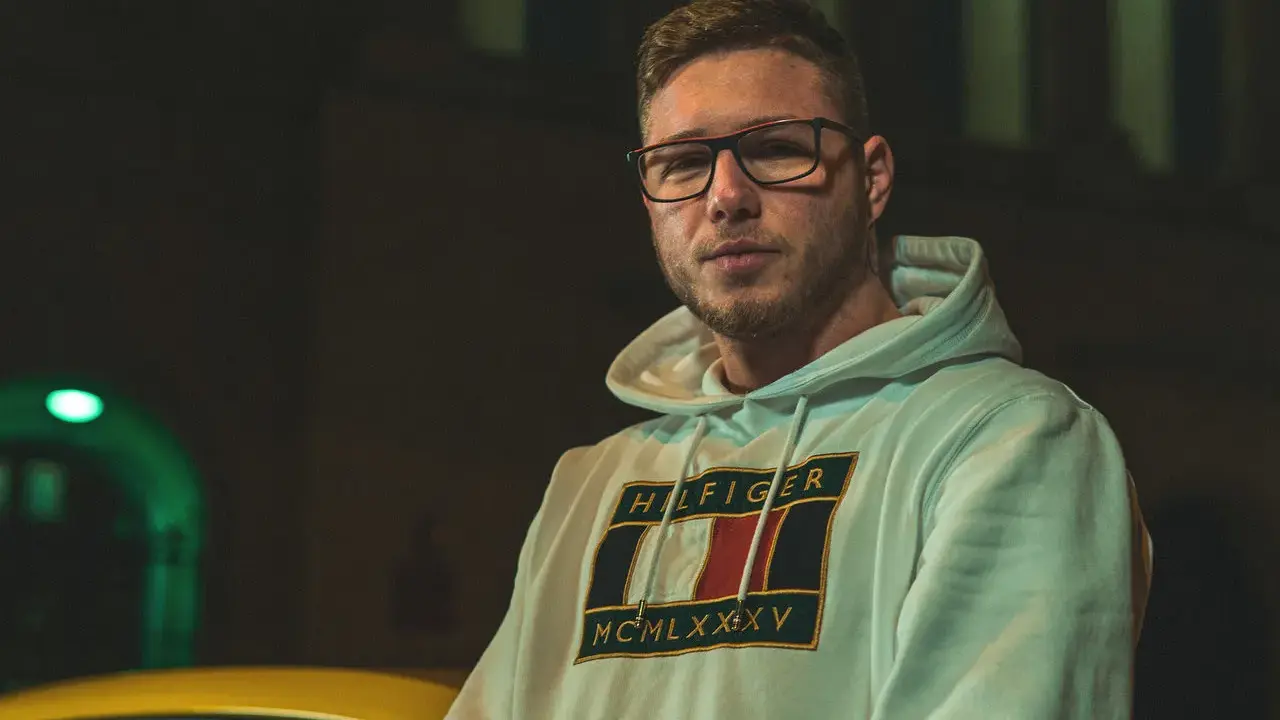 Kubańczyk cały ja utwory. Młody mężczyzna w okularach i bluzie z kapturem Tommy Hilfiger, nocą, obok żółtego samochodu.