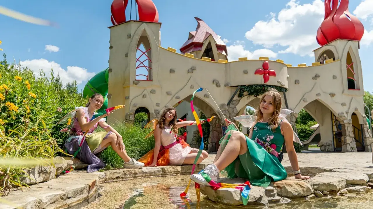 Trzy dziewczynki w strojach wróżek siedzą nad wodą przed bajkowym zamkiem. To jedne z wielu magiczne ogrody atrakcje.