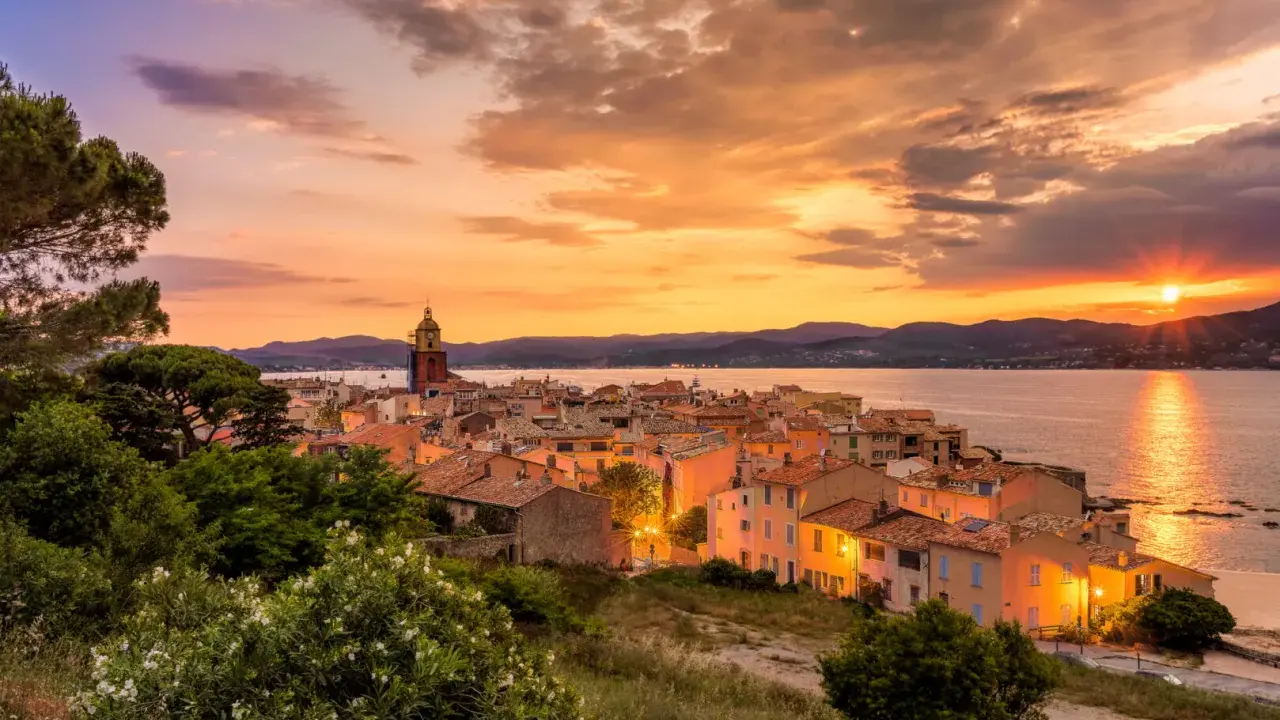 Sonnenuntergang über St. Tropez. Die Sonne versinkt im Meer, beleuchtet die Stadt und den Kirchturm in warmen Farben.