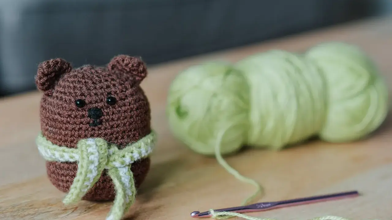 Os **sonajeros de crochet patrones gratis** son adorables. Un osito marrón con bufanda verde y blanca, junto a ovillos de lana y una aguja de crochet.