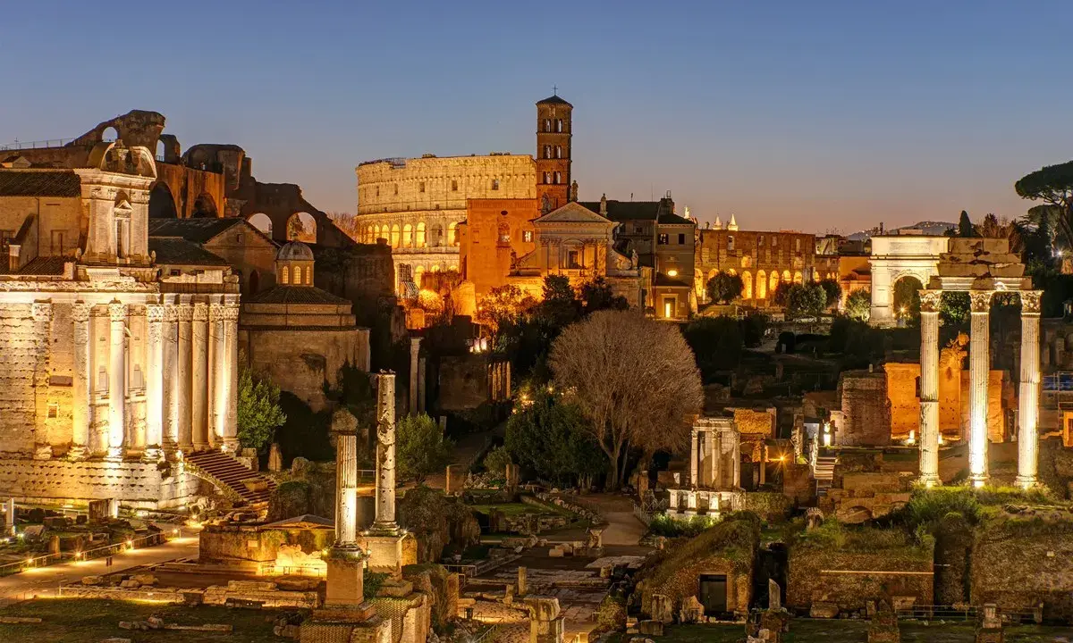 Antike Ruinen des Forum Romanum in Rom bei Nacht. Der Kolosseum und ein Triumphbogen sind beleuchtet.