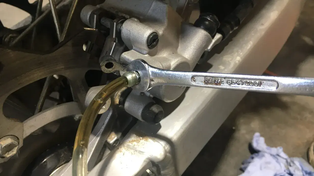 Klucz nasadowy odkręca odpowietrznik zacisku hamulcowego motocykla, podczas gdy przez przezroczysty wężyk płynie płyn. To etap wymiany płynu hamulcowego.