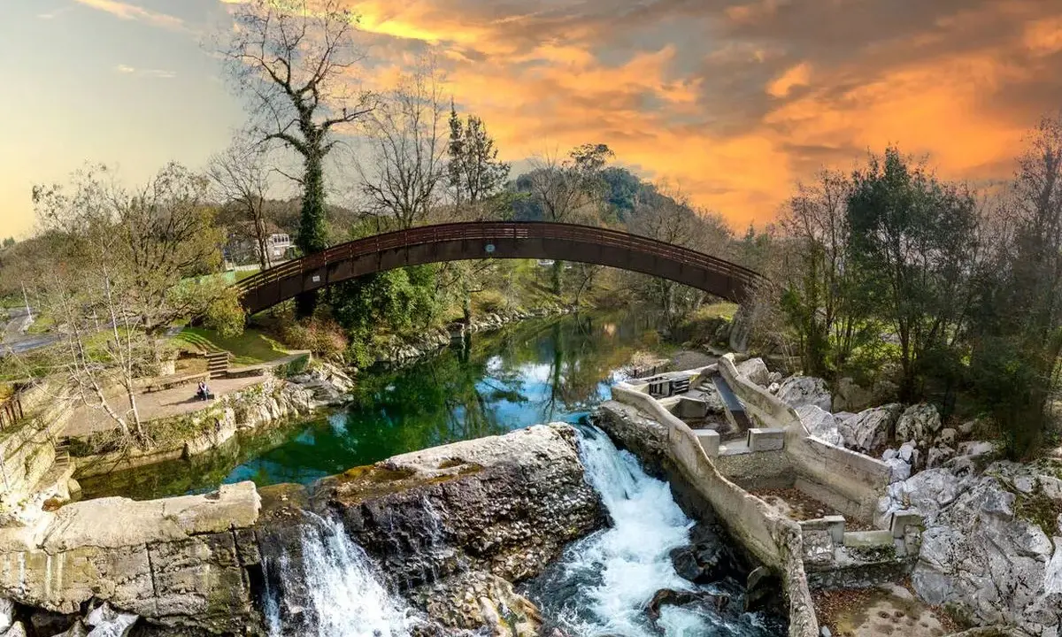 Un puente arqueado cruza un río verde, con cascadas y ruinas de piedra. Un paisaje idílico que invita a descubrir pueblos bonitos cerca de Puente Viesgo.