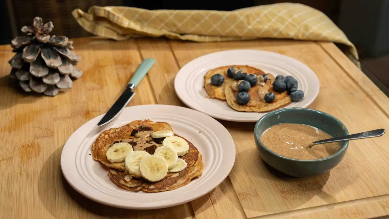 Pancakes ig bas farine complète, garnis de bananes, de chocolat et de myrtilles, avec un bol de beurre d'amande.