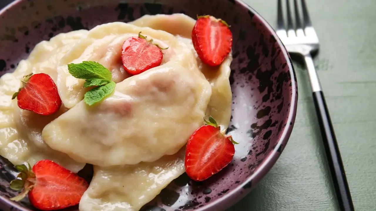 Pyszne pierogi z truskawkami, udekorowane świeżymi owocami i miętą, czekają w misce na widelcu.