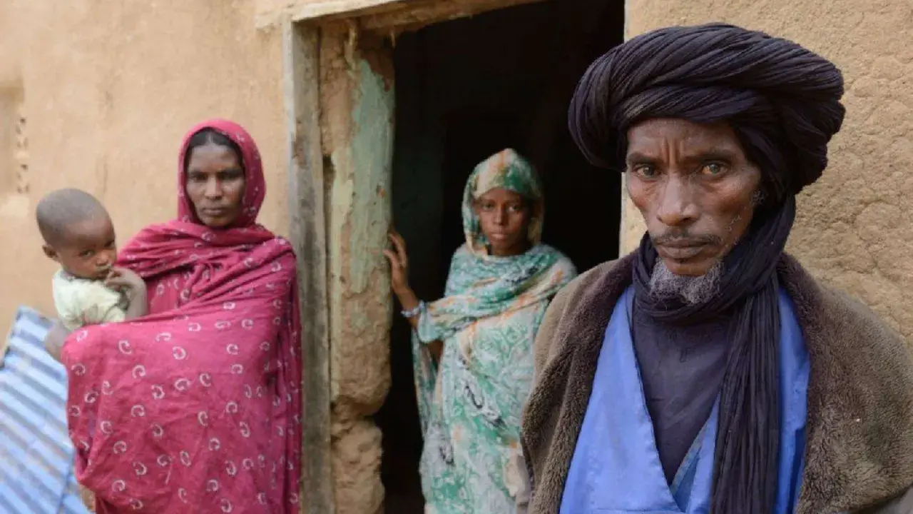 Ein Tuareg-Mann mit schwarzem Turban und ein Kind mit seiner Mutter vor einem Lehmhaus in der Sahara.