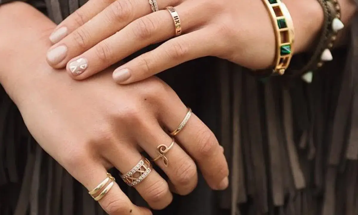 Mani adornate con anelli, tra cui uno con numeri romani, un bracciale colorato e un anello a forma di "S". Non c'è un anello da single specifico, ma la varietà suggerisce stile personale.