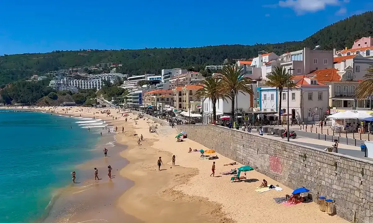 Sonne, Strand und Meer in Sesimbra, Portugal. Menschen genießen den Tag am goldenen Sandstrand, während im Hintergrund die Stadt und grüne Hügel aufragen.