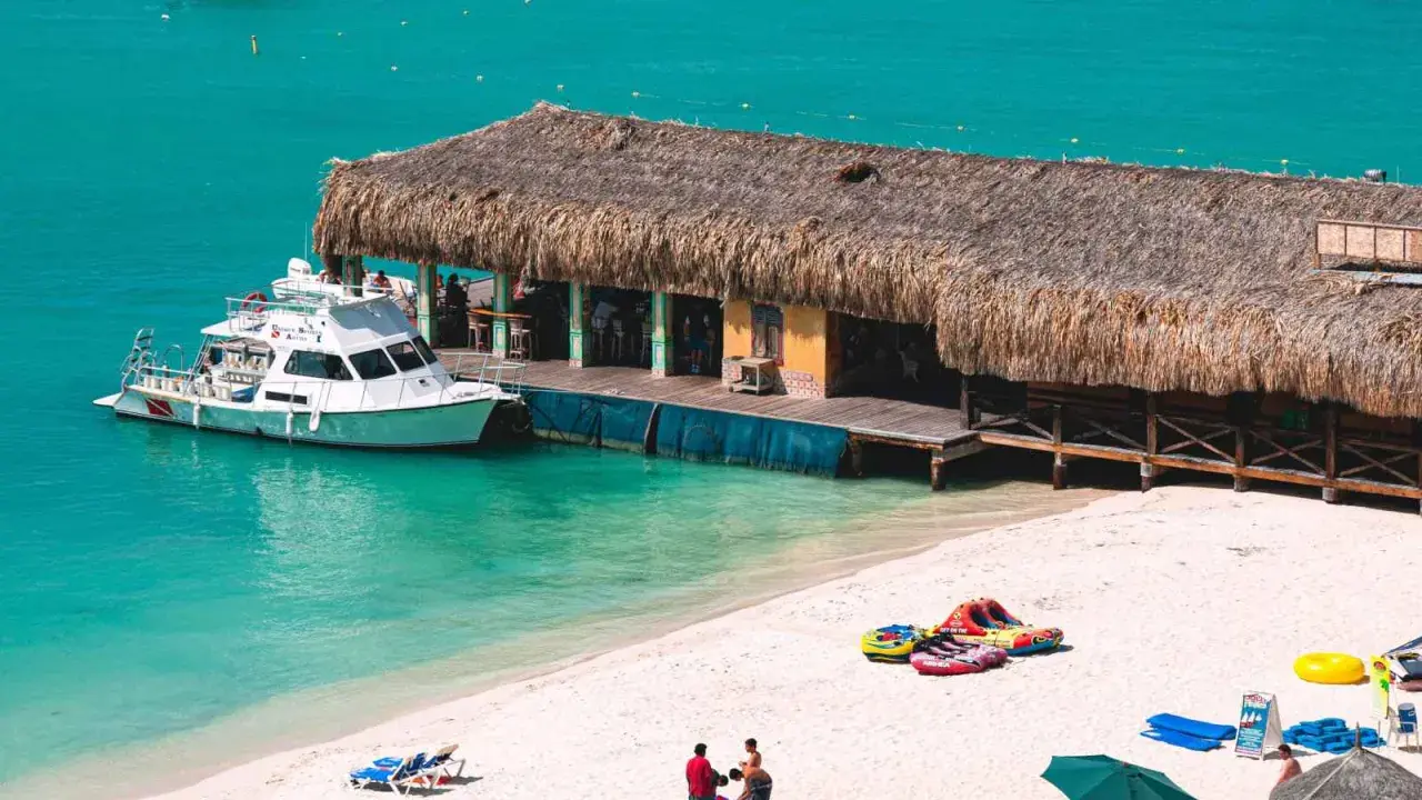 A boat docks at a thatched-roof beach bar on a sunny day. This scene embodies the welcoming spirit of Aruba, a destination known for being gay friendly.