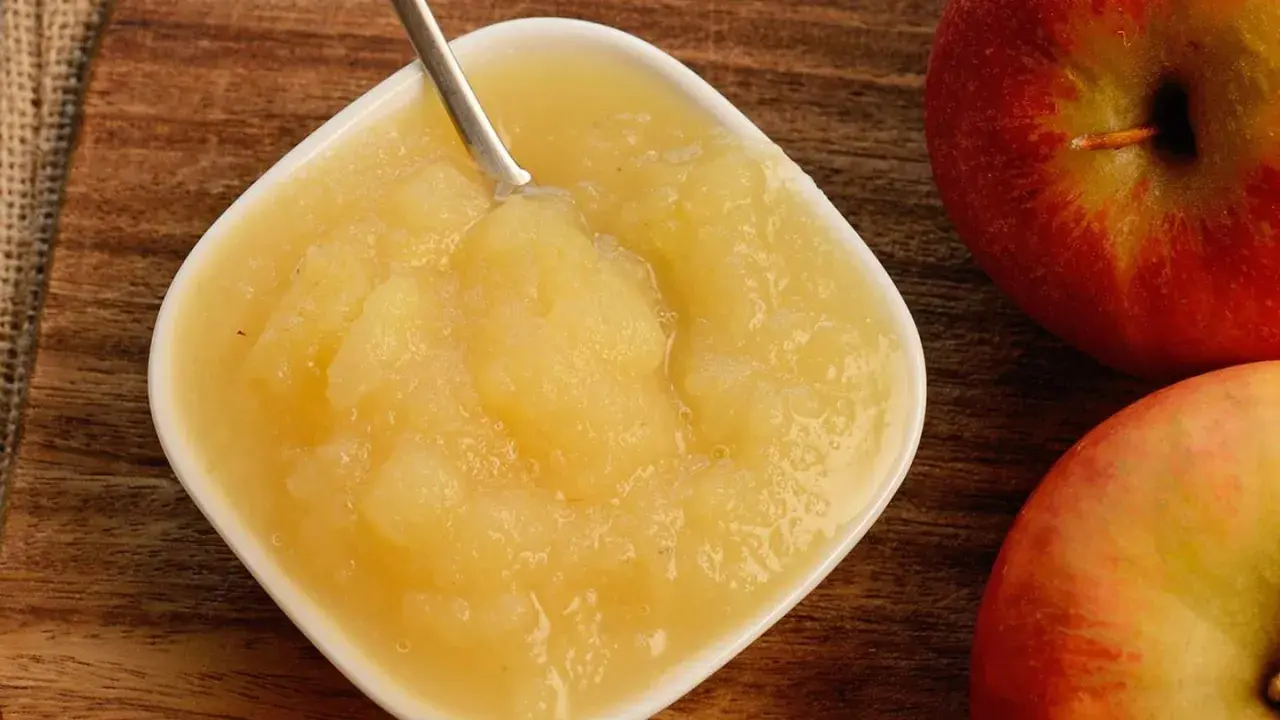 Frisches Apfelmus mit Schale, zubereitet im Thermomix, in einer weißen Schale auf Holzuntergrund. Daneben liegen zwei rote Äpfel.