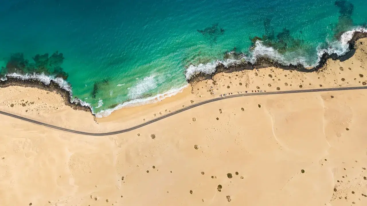Luftaufnahme eines einsamen **Surferstrands** auf **Fuerteventura**. Türkisblaues Meer trifft auf goldgelben Sand und eine felsige Küste. Eine Straße schlängelt sich entlang der Küste.
