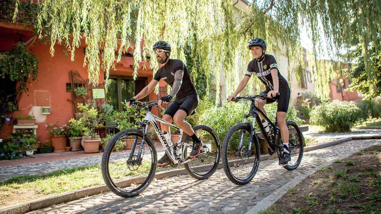 Coppia in bici da mtb a Rimini, pedalando su un sentiero acciottolato tra case e vegetazione rigogliosa.